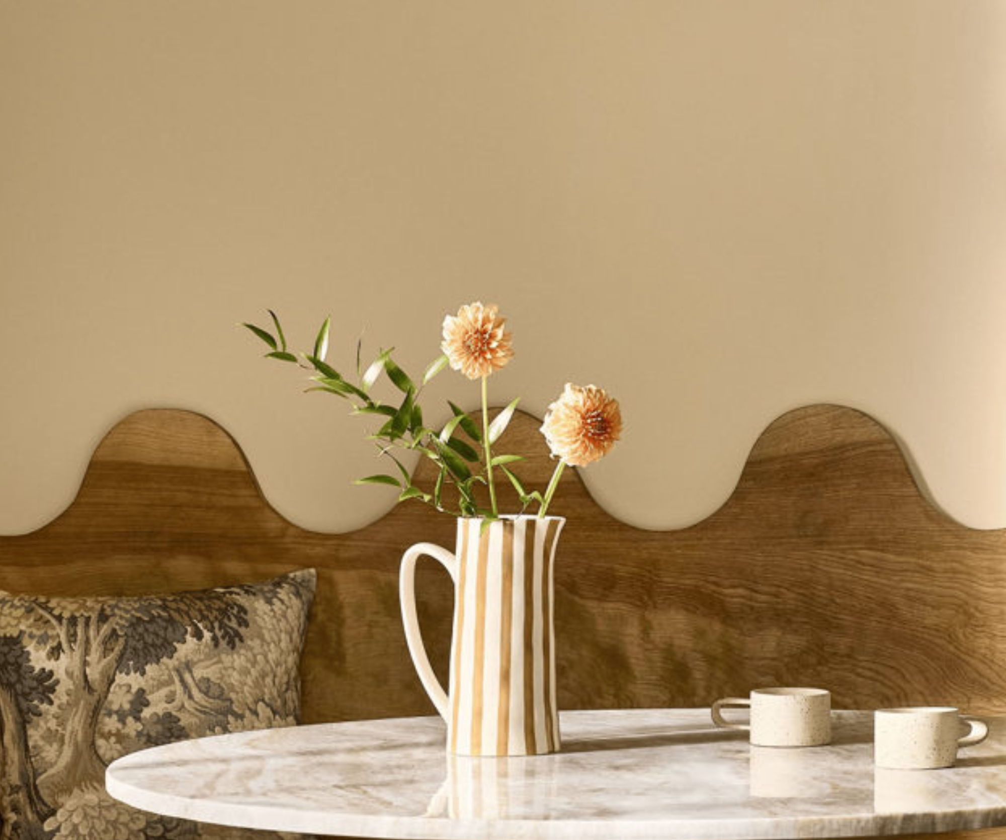 A close-up of a marble tabletop with a striped ceramic pitcher holding orange flowers, positioned in front of a wooden booth with a distinct wavy backrest.