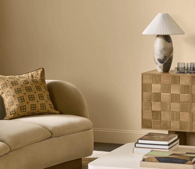 A cozy living room corner showing a section of a cream couch with a patterned pillow, a white coffee table with books, and a textured wooden cabinet topped with a pleated lamp.